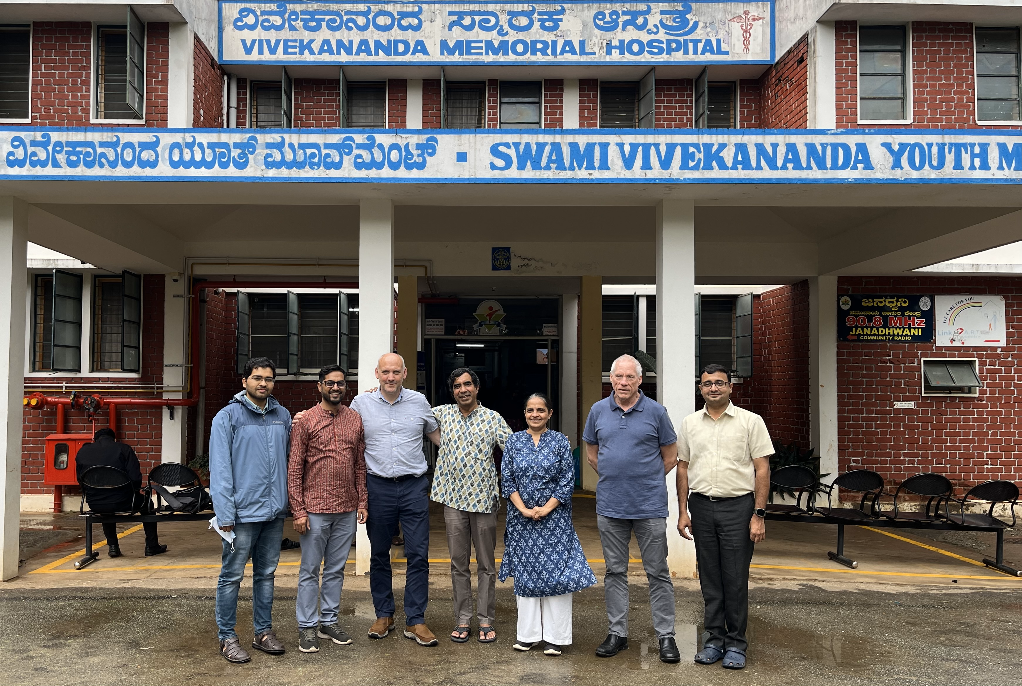 6 men and 1 woman standing in front of a building in a row and smiling